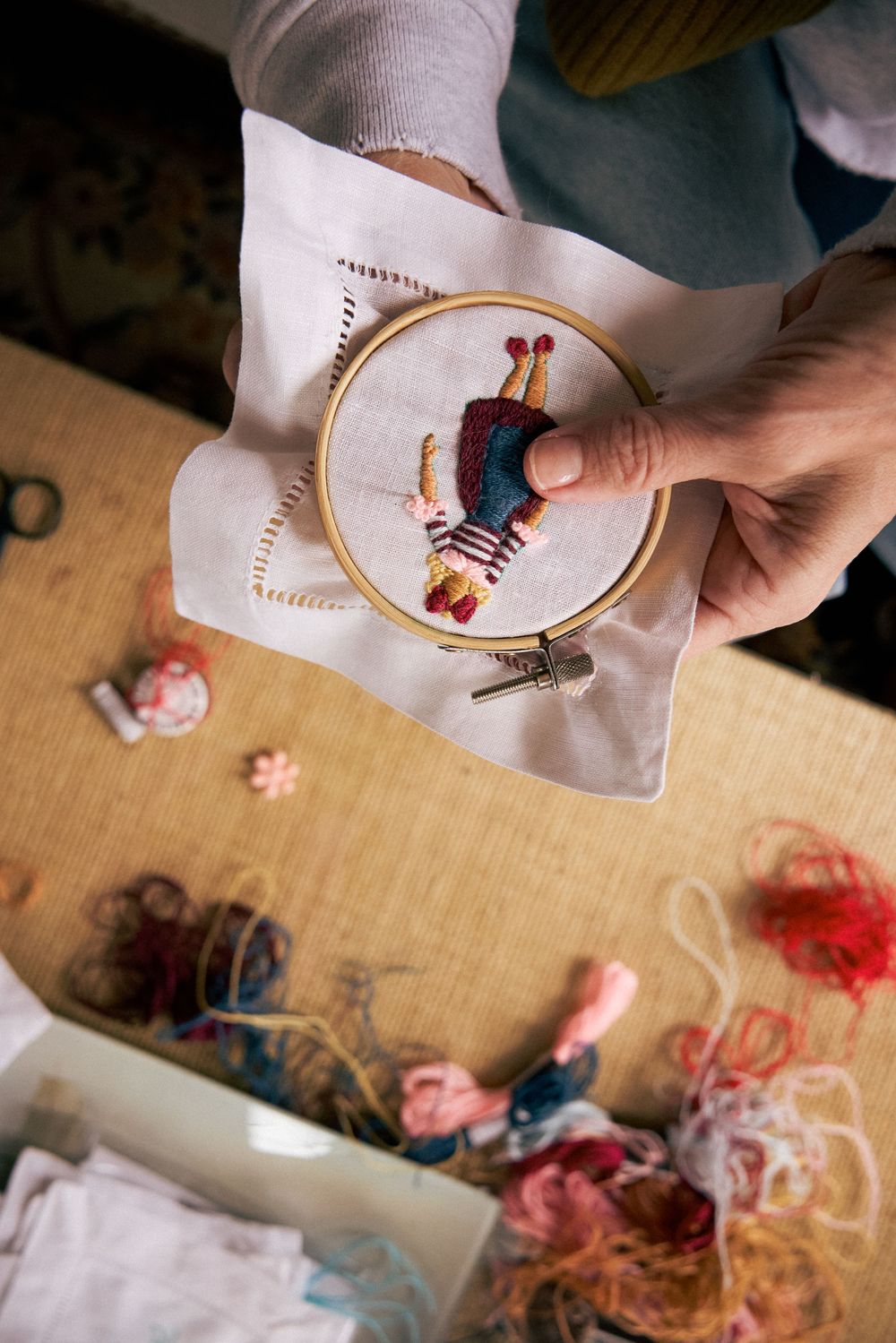 Hands holding an embroidery hoop with a colorful stitched figure above a table scattered with colorful threads.
