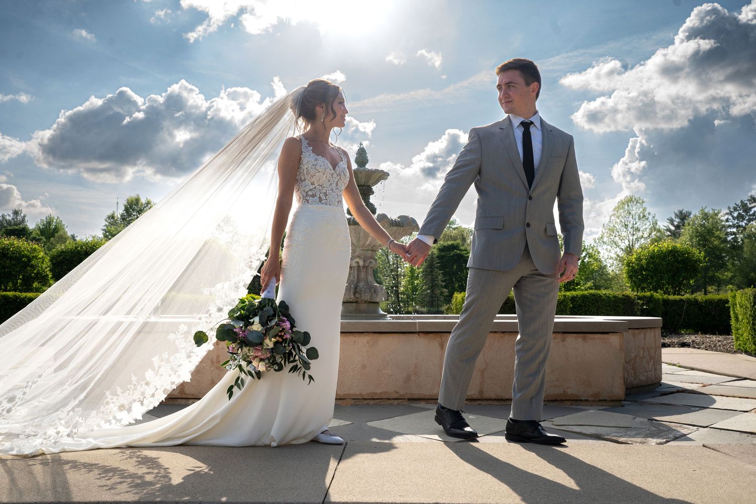 Bride and groom hold hands outdoors at elegant garden venue, bride in white gown with veil, groom in gray suit.