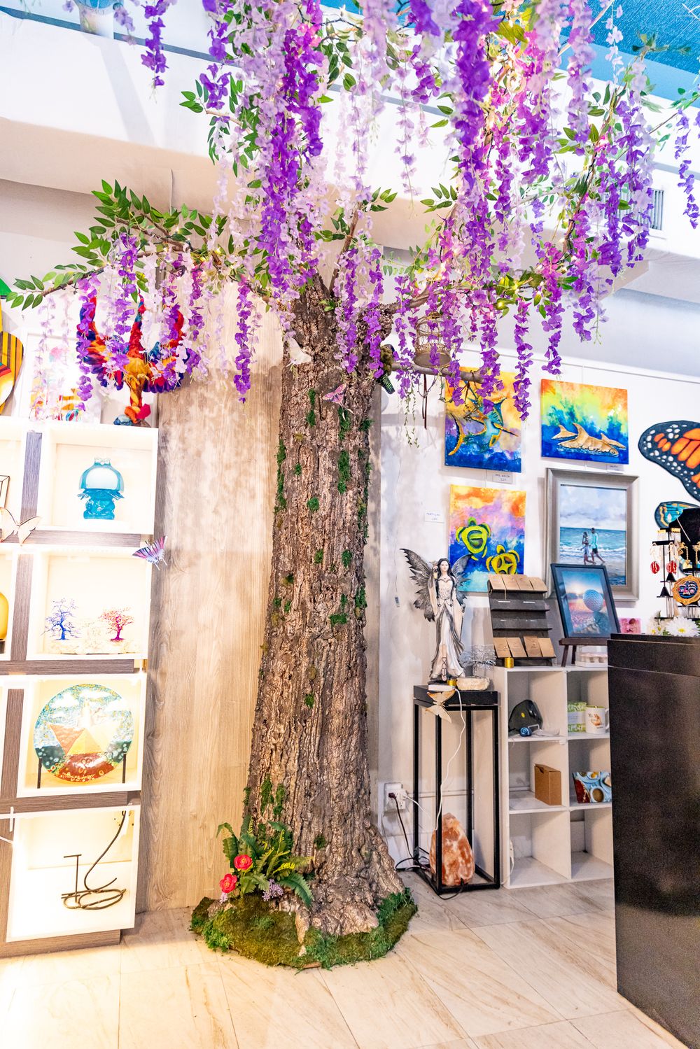 Art gallery interior with decorative wisteria tree featuring purple blooms, surrounded by colorful paintings and sculptures.