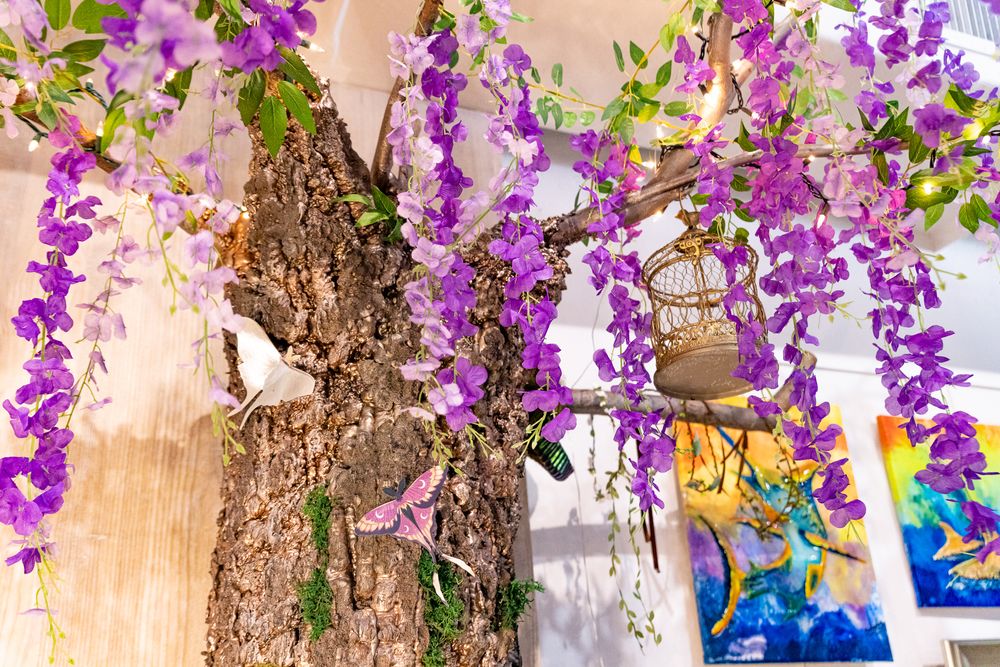 A decorative tree adorned with cascading purple wisteria flowers and a hanging birdcage in an indoor setting.