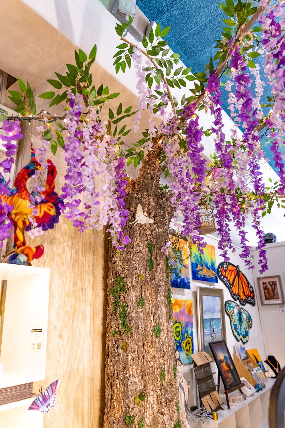 Decorative indoor tree with hanging purple wisteria flowers, butterfly art, and colorful paintings in a gallery shop.