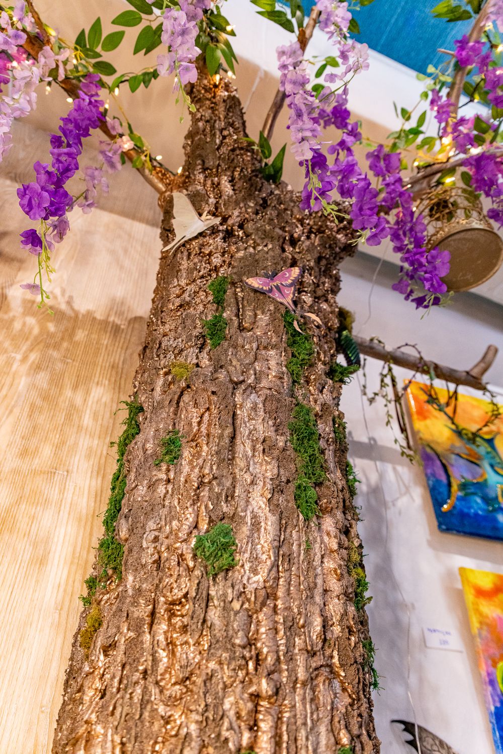 A tree trunk adorned with moss and purple wisteria flowers displayed indoors in an art gallery setting.