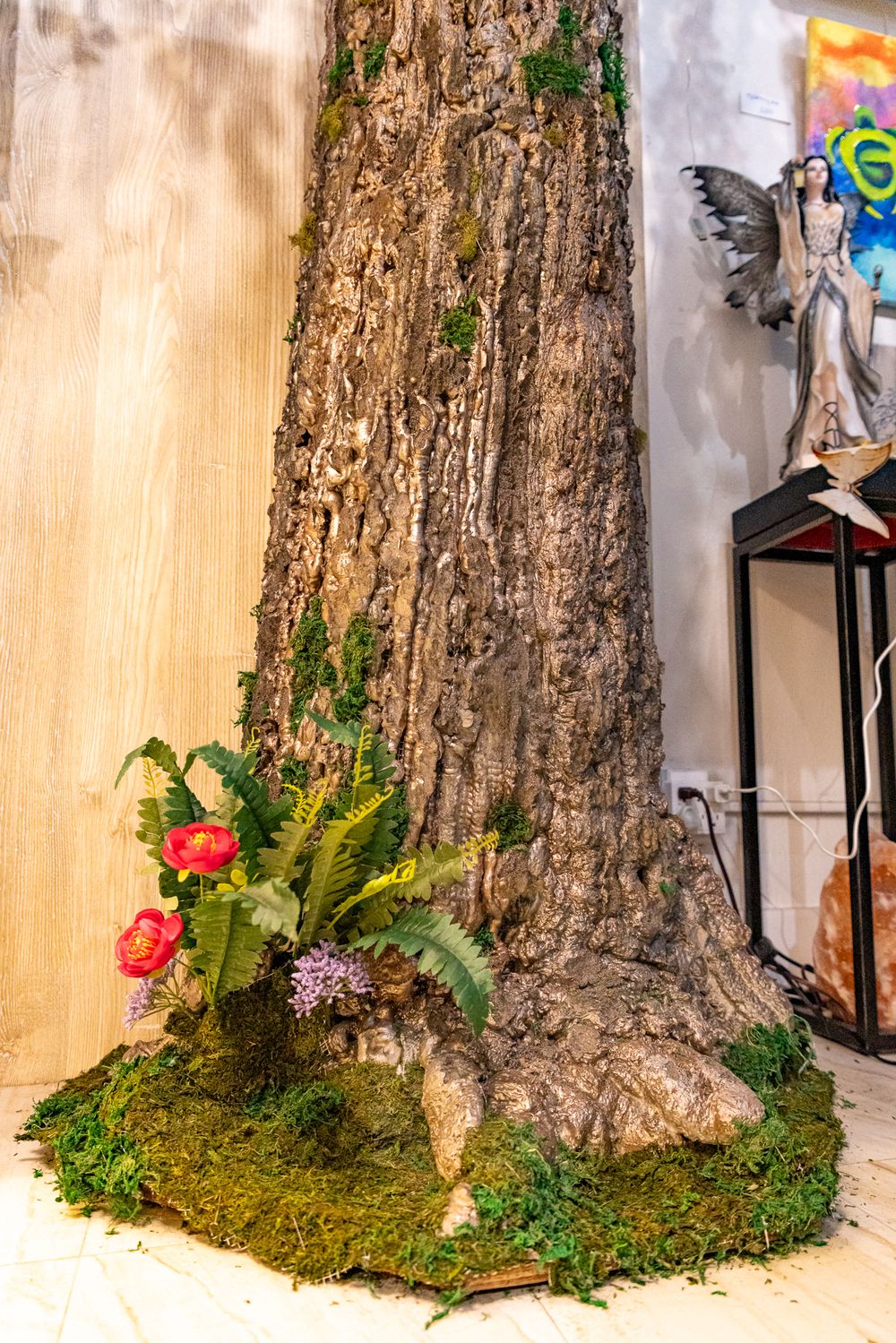 Large tree trunk display with moss, ferns, and red flowers at base, fairy statue visible in background.