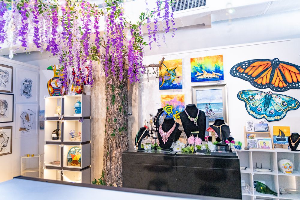 Colorful art gallery with butterfly paintings, jewelry display, and hanging purple wisteria flowers in a bright white boutique shop.