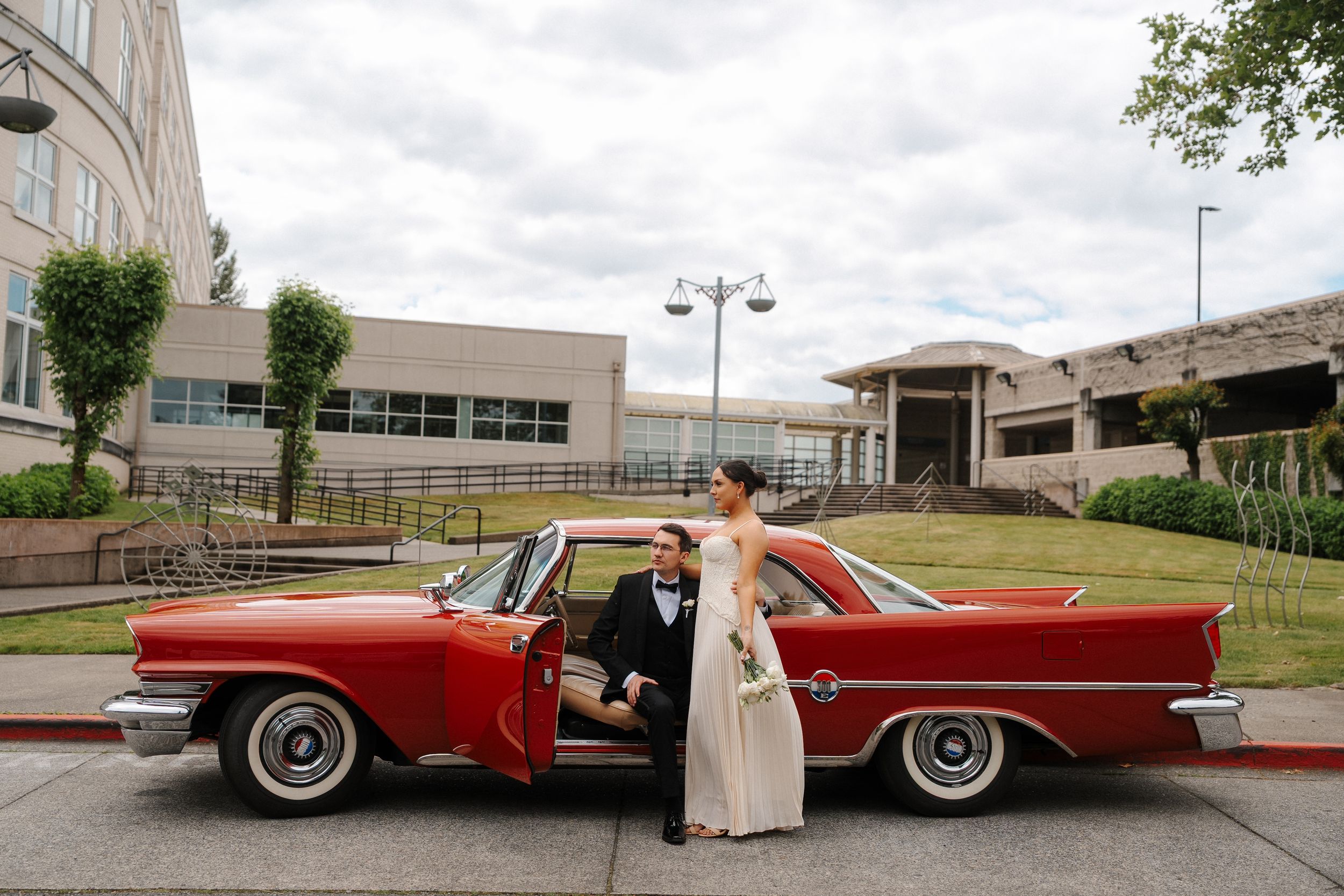 Bride and groom posing beside a classic red vintage car on their wedding day outside a modern building.