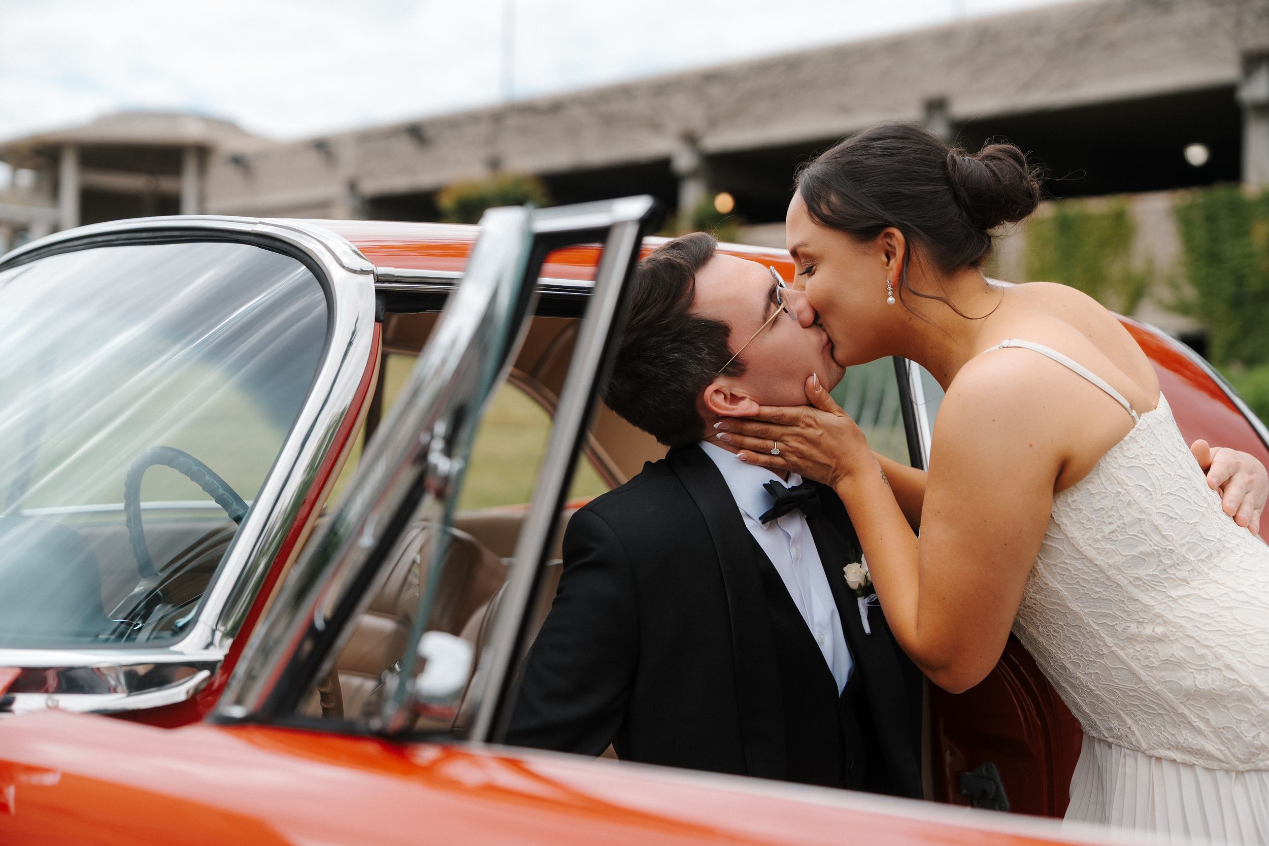 Bride and groom share a romantic kiss beside a vintage orange convertible car on their wedding day.