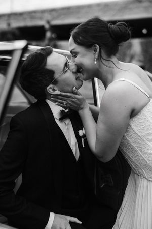 Black and white photo of a bride and groom sharing a romantic kiss near a vintage car on their wedding day.