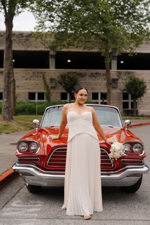 Bride in white gown holding bouquet poses in front of a classic red convertible car outdoors.