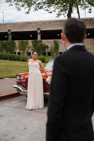 Young couple in formal wear poses by a vintage red classic car in a parking lot, woman in white gown smiling.