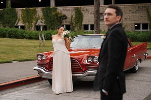 Bride in white gown holds bouquet near red classic car as groom in black suit stands in foreground.