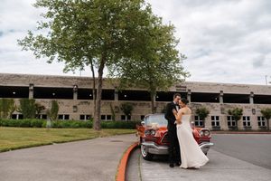Bride and groom posing beside a vintage red car in front of a large building with a tree backdrop.