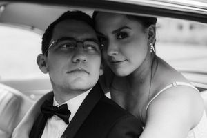 Black and white wedding photo of a couple in formal attire sitting in a vintage car, groom in tuxedo and bride in white dress.