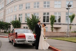 Bride and groom pose beside a vintage red classic car outside an elegant building on their wedding day.