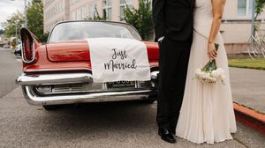 Newlywed couple standing beside a classic red car with a Just Married banner at their wedding.