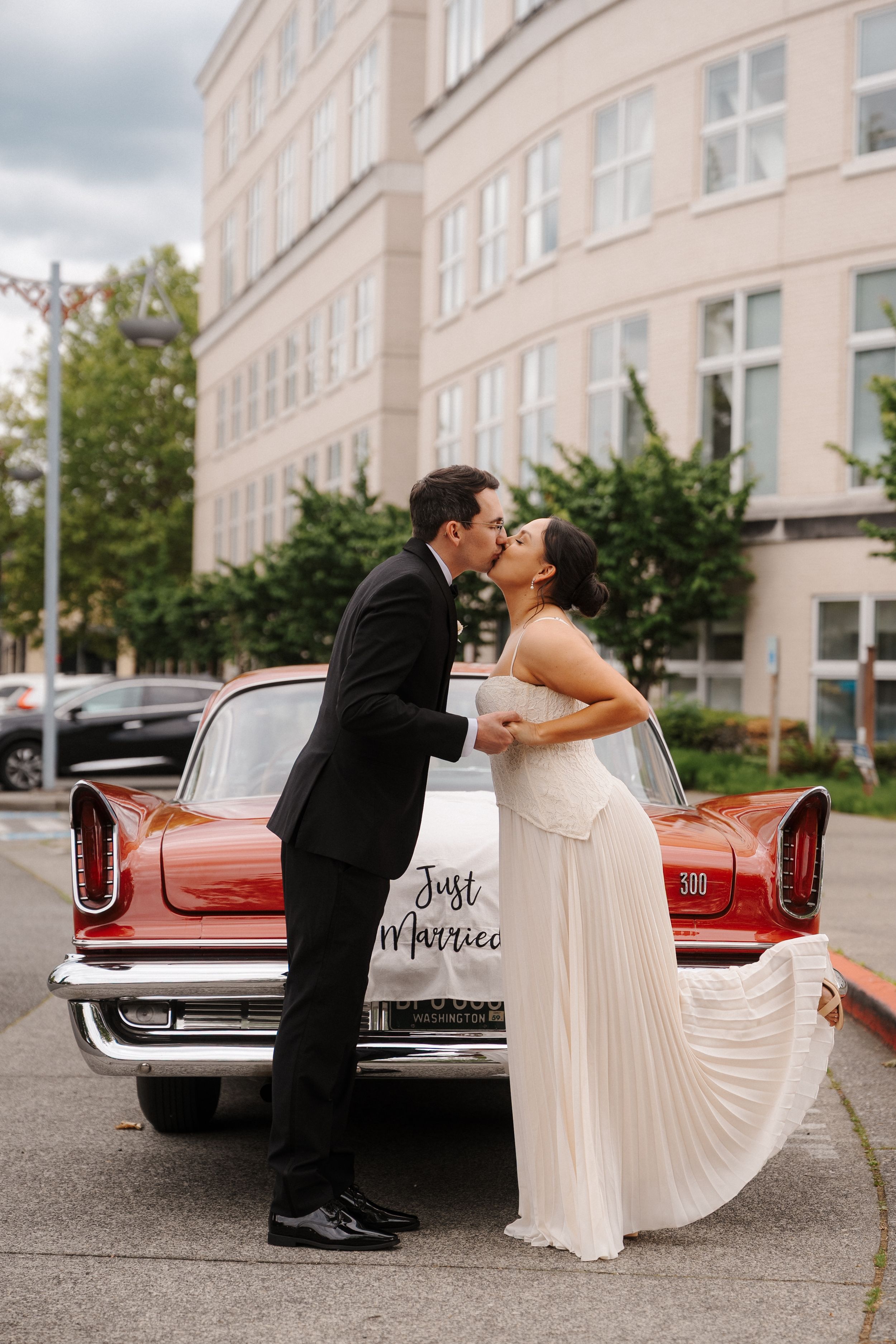 Newlyweds share a kiss leaning against a red classic car with a Just Married sign outside a building.