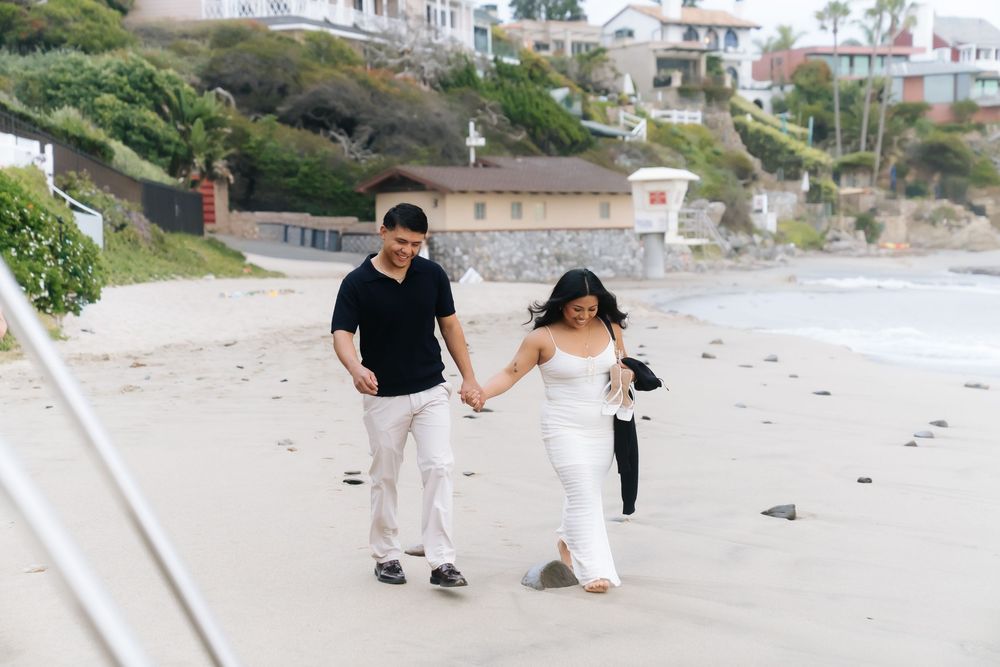 Couple holding hands walking on a sandy beach with coastal homes in the background on a cloudy day.