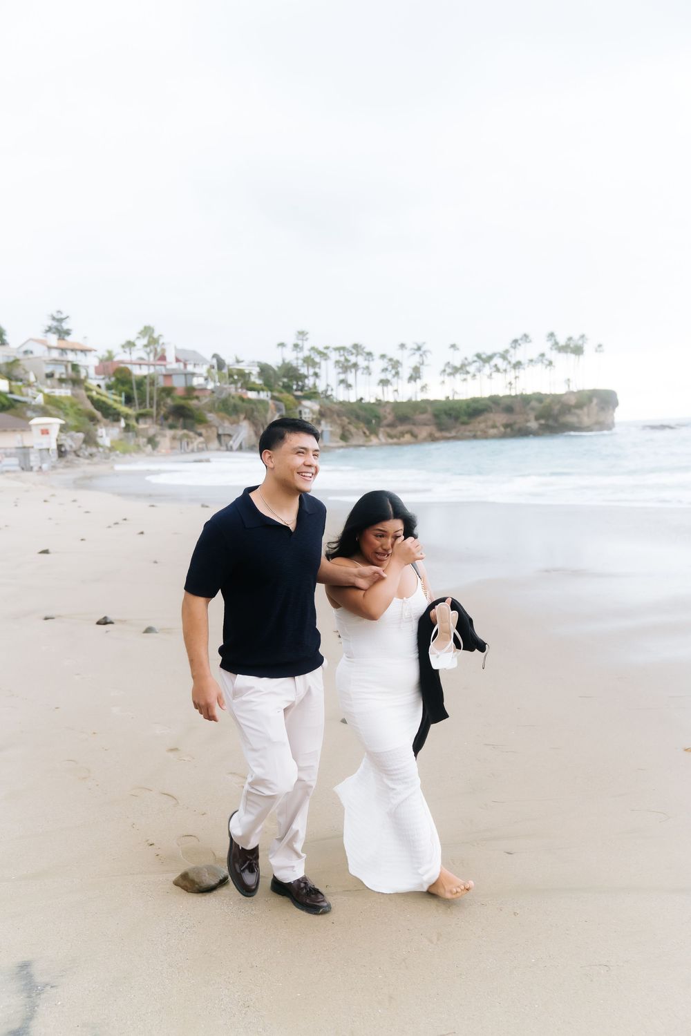 Couple walking on beach, man in black shirt and white pants, woman in white dress, coastal cliffs in background.