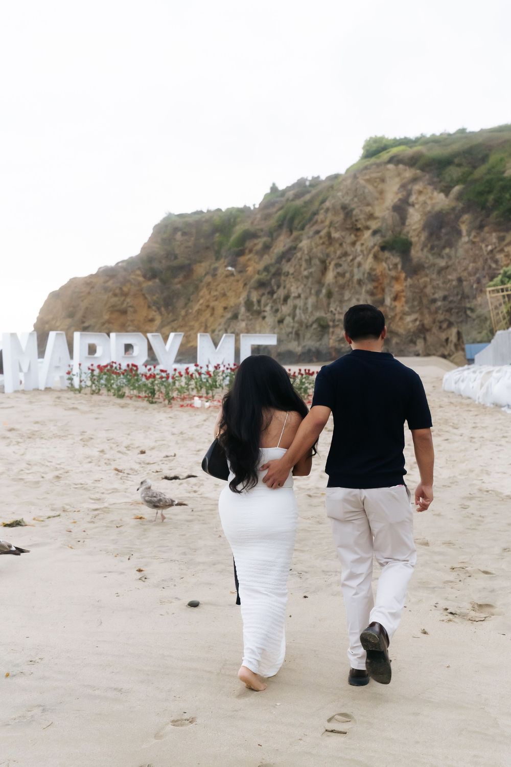 Couple walking hand-in-hand on beach toward large MARRY ME sign with flowers and cliffs in background.