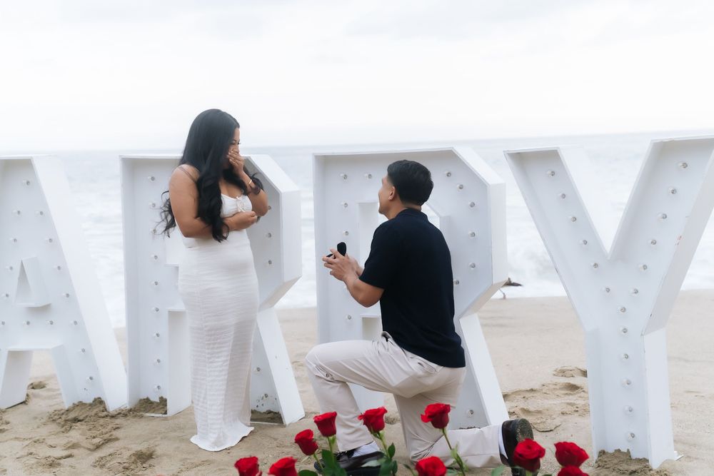 Man proposing on beach with ring, woman in white dress, large HAPPY letters and red roses.