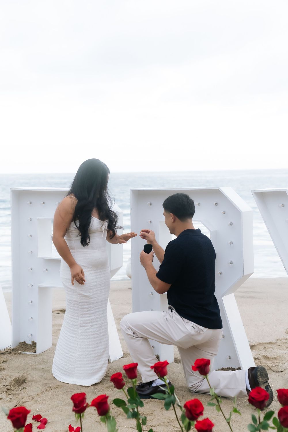 Man proposes to woman on beach with red roses and white letters, holding ring box while kneeling.