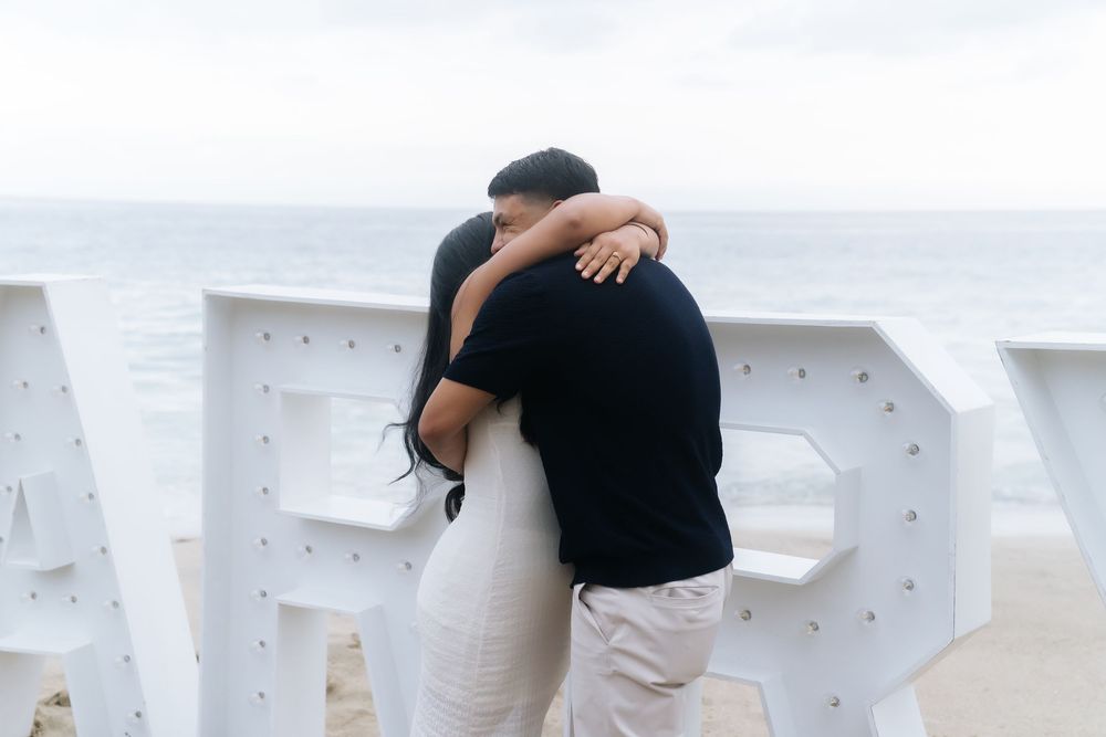 Couple embracing in front of large white marquee letters spelling MARRY on a beach with ocean views.