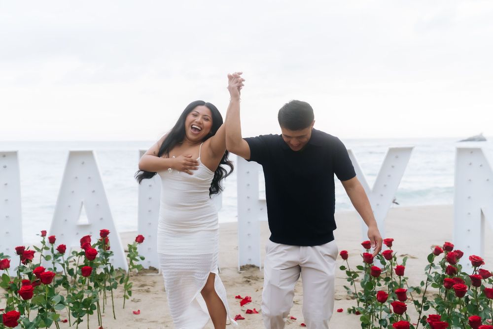 Couple celebrating beach proposal with red roses and marquee letters, woman in white dress raising hands joyfully.