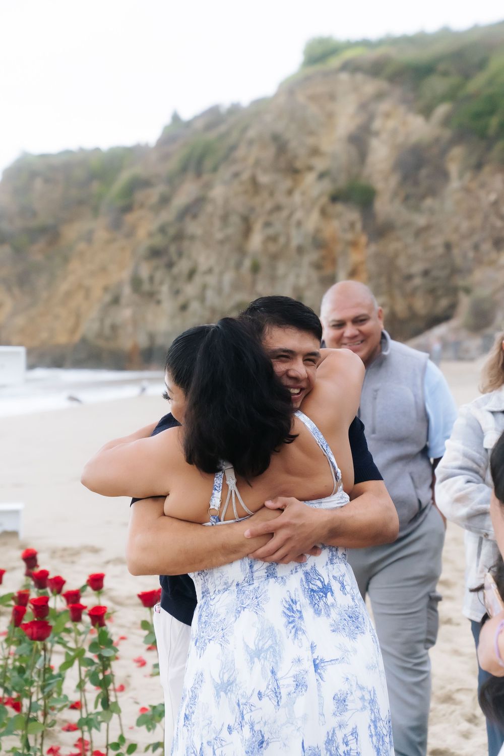 Couple embracing on a beach with red roses, smiling man hugging woman in floral dress near rocky cliffs.