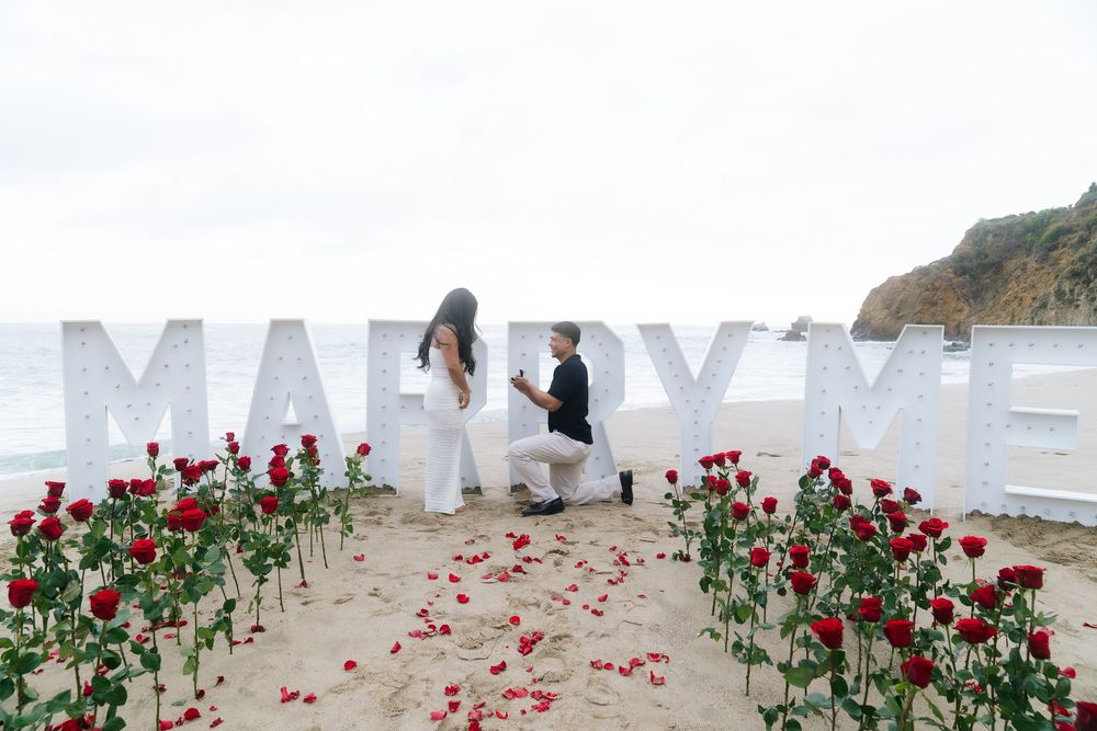Man proposes to woman on beach with red roses and large MARRY ME marquee letters by the ocean.