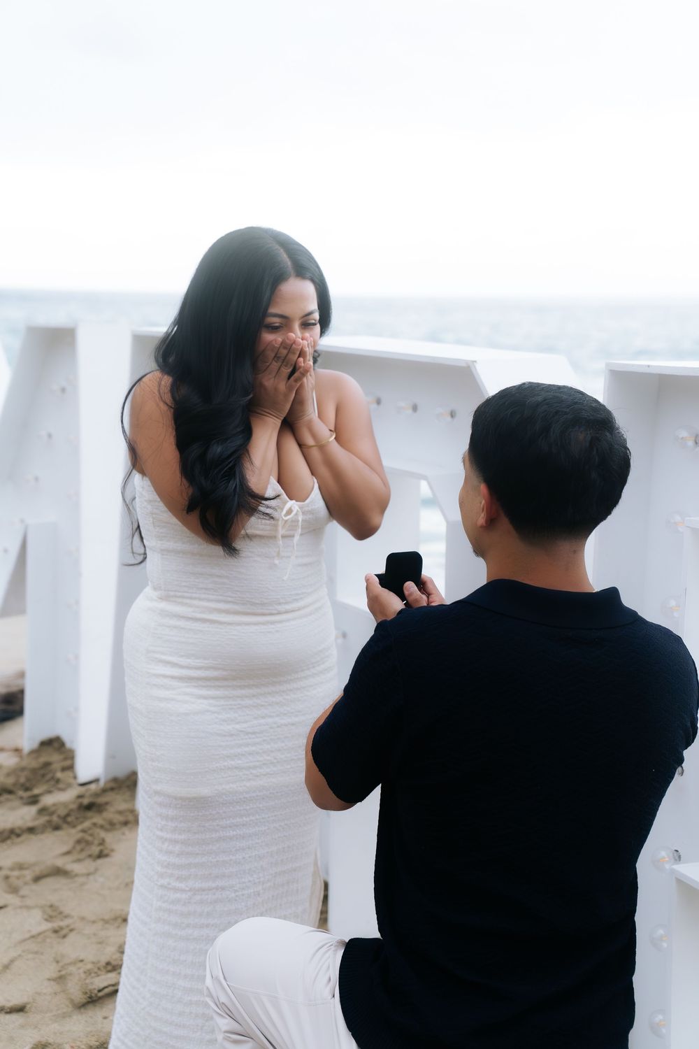 A man kneeling on a beach proposes with a ring box to a surprised woman in a white dress covering her mouth.