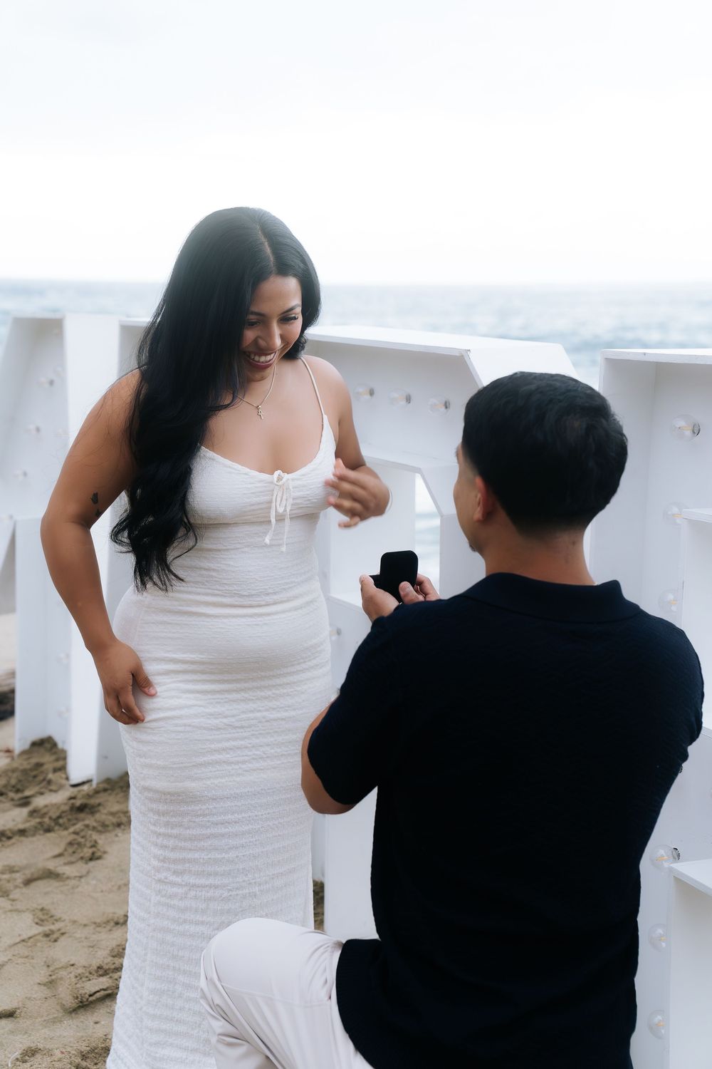 Man in black kneeling to propose to woman in white dress on sandy beach with white chairs.