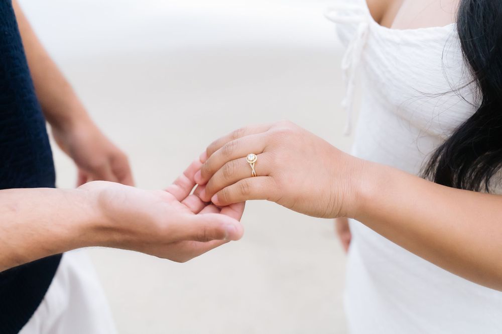 Close-up of couple holding hands with a diamond engagement ring on the woman's finger on a beach.