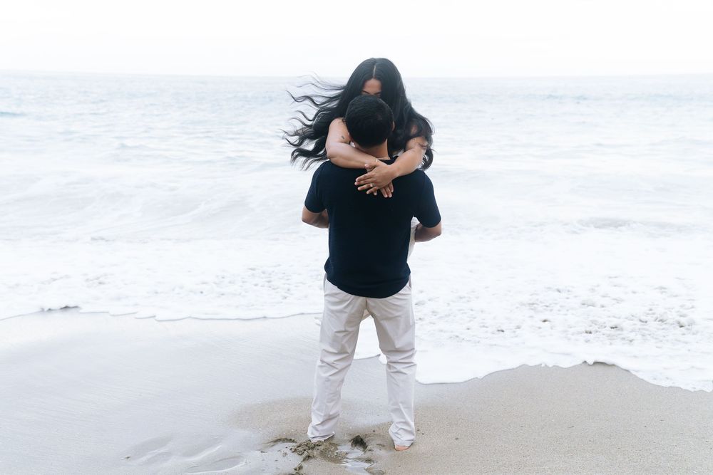Couple embracing on a sandy beach, woman wrapped around man's back with ocean waves in the background.