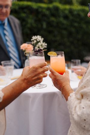 Two people clinking pink cocktail glasses at an outdoor wedding reception with white tablecloth and greenery.