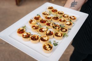 Server holding a white platter of mini tartlets filled with savory toppings and fresh herbs at a catered event.
