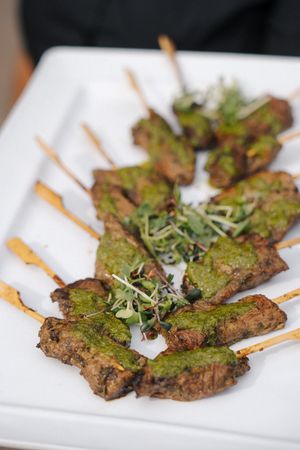 Grilled meat skewers topped with green herb sauce and microgreens, served on a white rectangular plate.