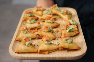 Golden fried pastry bites topped with fresh guacamole and diced tomatoes served on a wooden board.