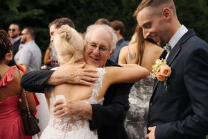 Elderly man warmly hugging bride in lace wedding dress while groom in navy suit smiles at outdoor wedding reception.