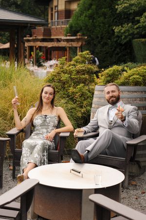 Elegant couple in formal attire relaxing outdoors at a garden venue, holding drinks and smiling at camera.