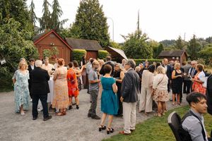 Guests mingle outdoors at a wedding reception near rustic red barn buildings surrounded by lush green trees.