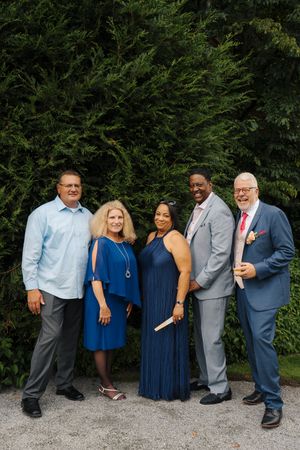 Five elegantly dressed guests pose together outdoors at a wedding, smiling in front of lush green trees.