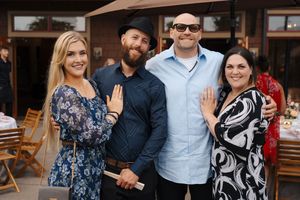 Two couples smile and show off engagement rings at an outdoor celebration venue.