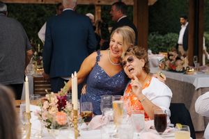 Two women laughing joyfully at a floral-decorated wedding reception table with candles and elegant glassware.