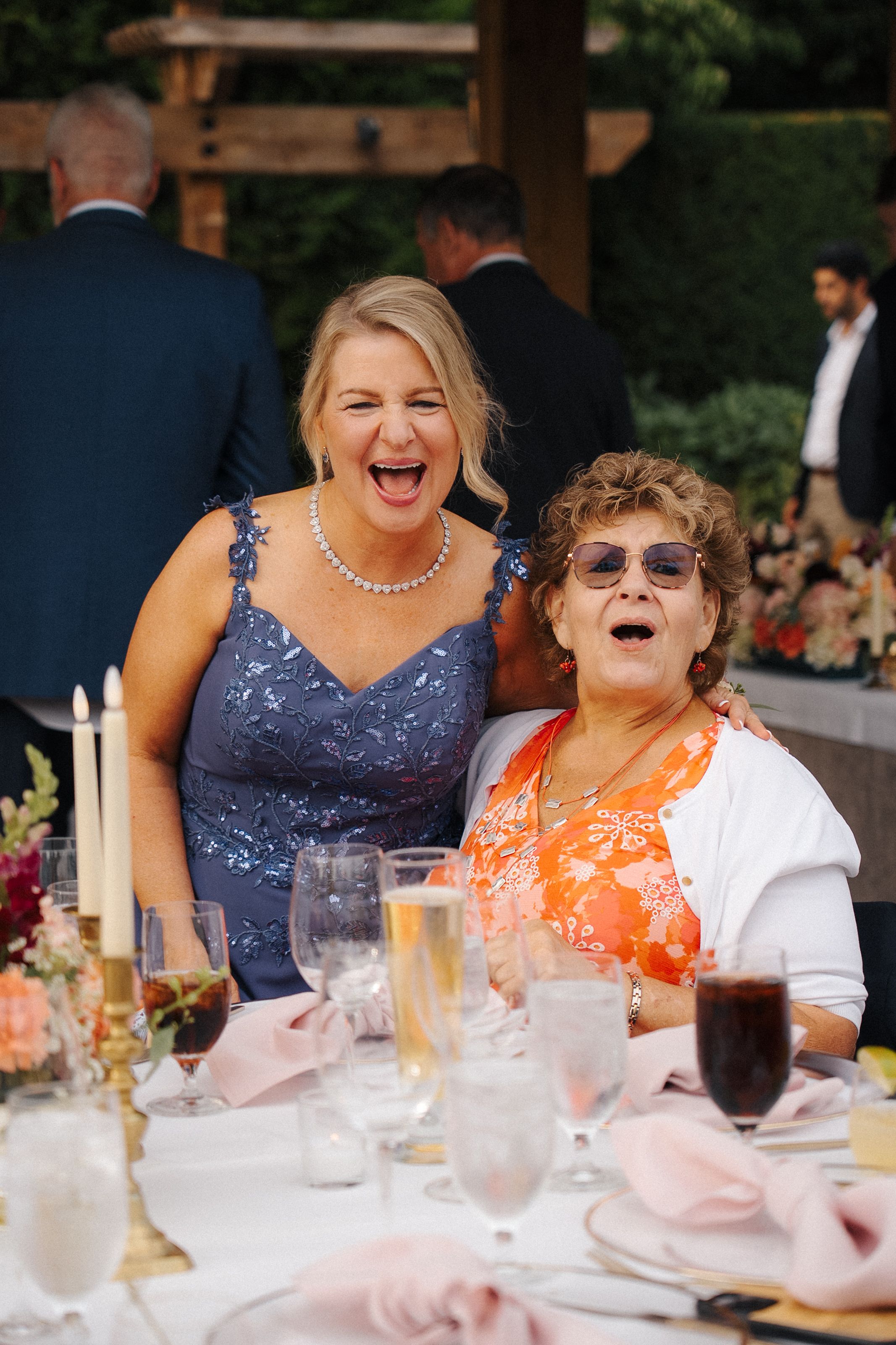 Two women laughing joyfully at a wedding reception table with elegant glassware and floral centerpieces.