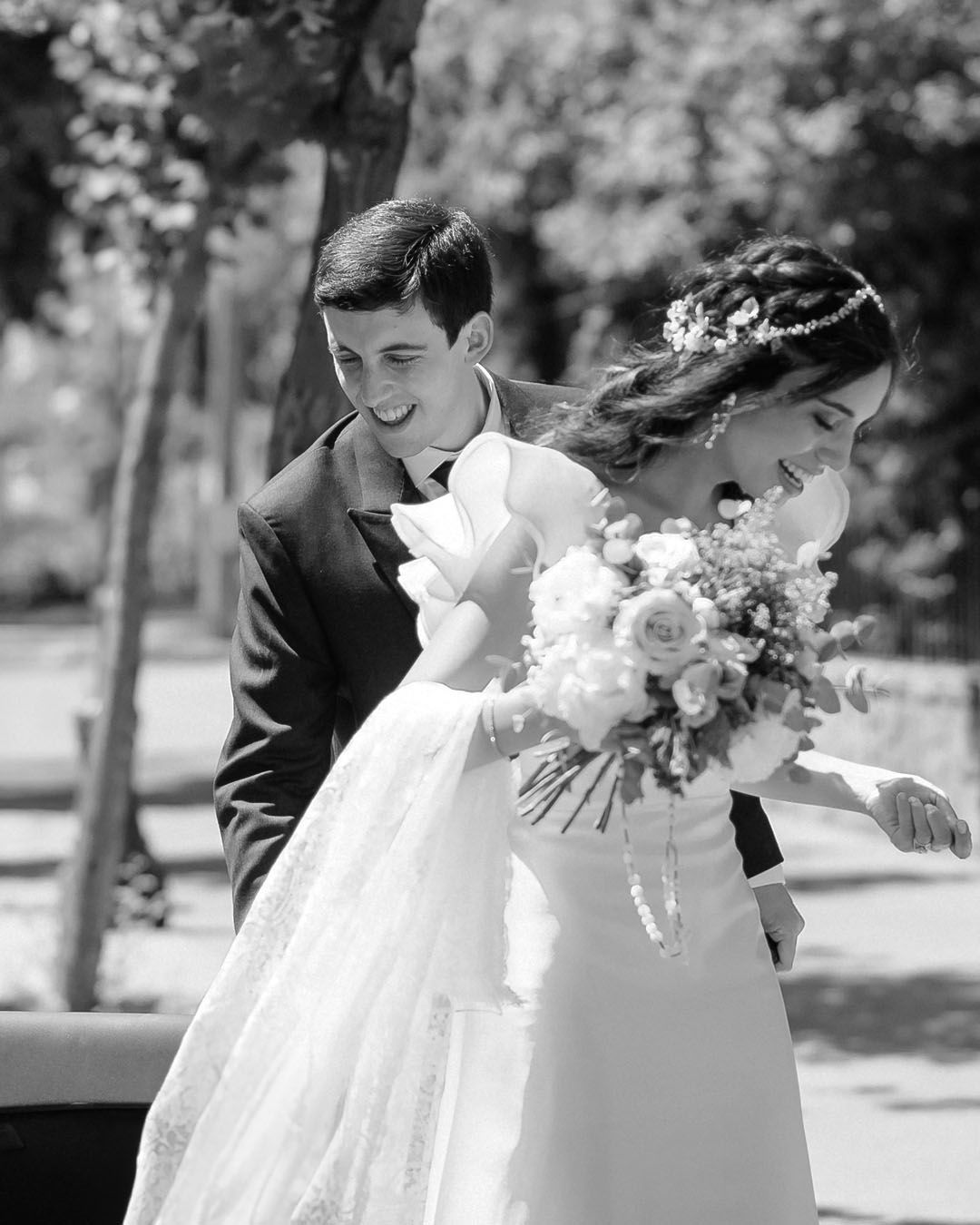 Black and white photo of laughing newlyweds walking together with bride holding a floral bouquet.