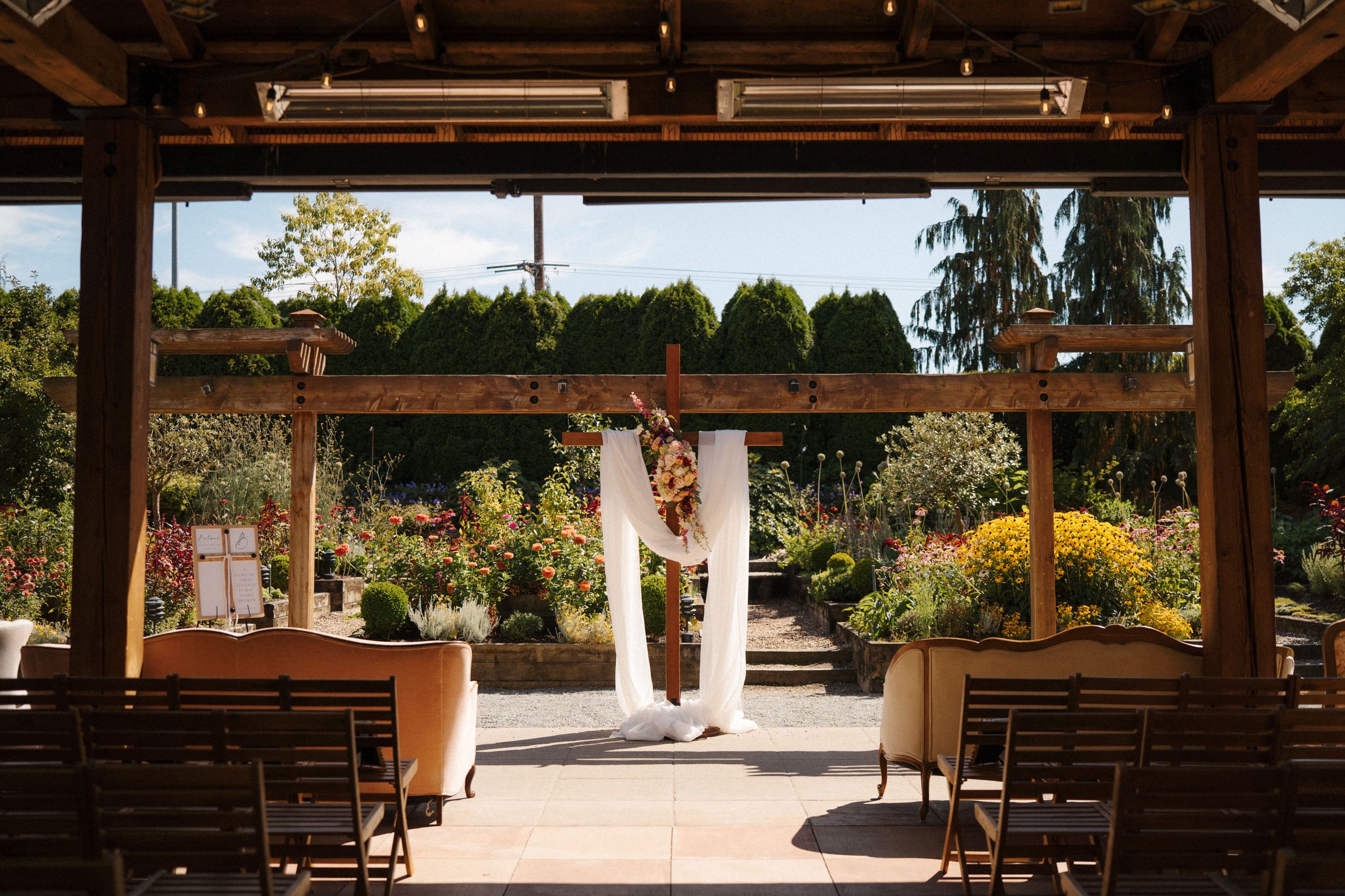 Elegant outdoor wedding ceremony setup under a wooden pergola surrounded by lush gardens and colorful flowers.