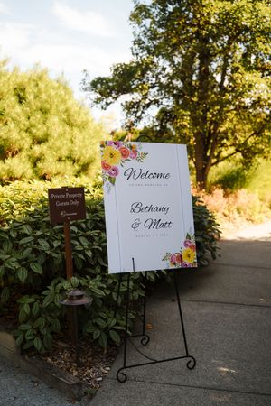 Floral wedding welcome sign for Bethany and Matt on an easel, surrounded by lush greenery outdoors.