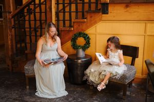 A bridesmaid and flower girl sit on benches reading books in a rustic wooden venue with a wreath decoration.