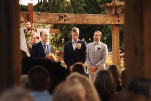 Groom and groomsmen standing at floral altar under wooden pergola during outdoor garden wedding ceremony.