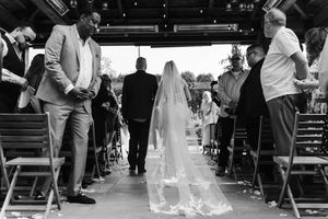 Black and white photo of a bride walking down the aisle in a flowing veil at an outdoor wedding ceremony.