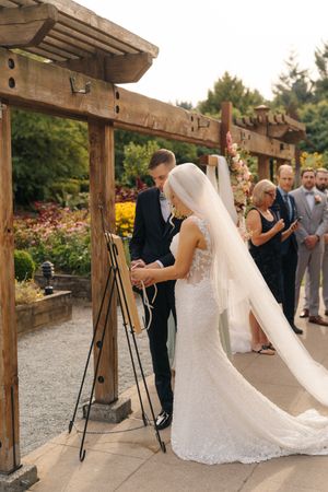 Bride and groom signing wedding documents under a wooden pergola surrounded by lush garden greenery.